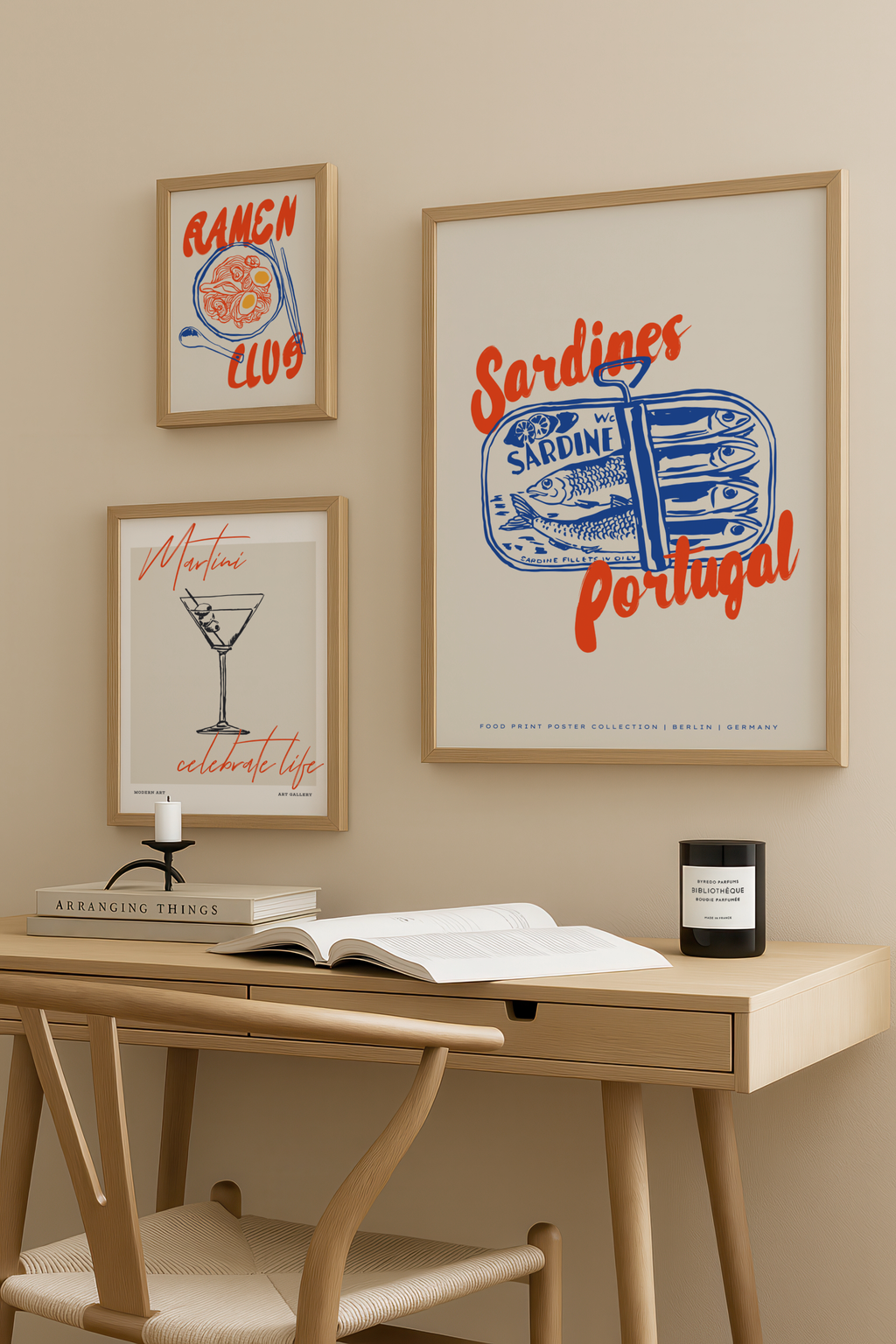 Ett litet träbord med en stol står mot en beige vägg dekorerad med Bozettos Bundle 3-pack Kökstavlor No. 1, med inramade bilder på sardiner, ramen och en martini. Ett ljus och en bok vilar på skrivbordet.