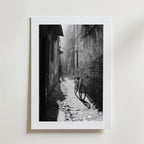 Quiet Echo Poster by Bozetto har ett svartvitt foto av en smal kullerstensgränd där solljuset kastar en cykels skugga genom dimman, tryckt på FSC-certifierat premiumpapper för en högkvalitativ finish.