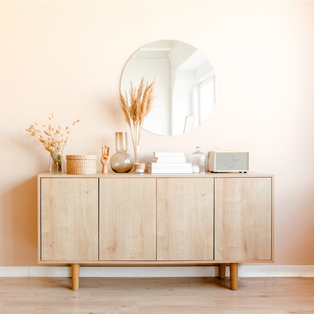 En sideboard i ljust trä med dekorativa vaser, böcker, en korg, torkat pampasgräs och en retroradio står mot en beige vägg, toppad med tidlös inredning och Bozetto Spegel Rund Cirkel 70 cm hängande ovanför.